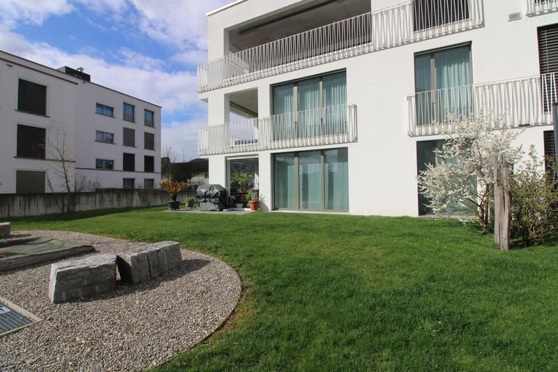 A well-maintained garden with a gravel pathway, a grassy area, and a stone bench. There are potted plants and a grill on the side. The garden is surrounded by a white building with glass windows and balconies.