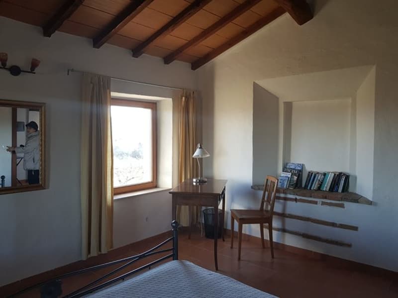Wooden ceiling beams, large window, wooden furniture including a desk and chair, shelves with books, and a bed frame.