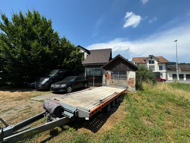 Two parked cars, a trailer, a small building, houses in the background, tree