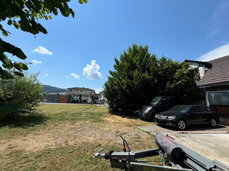 A wide lawn area in front of the house, two parked cars in the driveway, and a tree nearby. In the background, there is a building and a gas station.