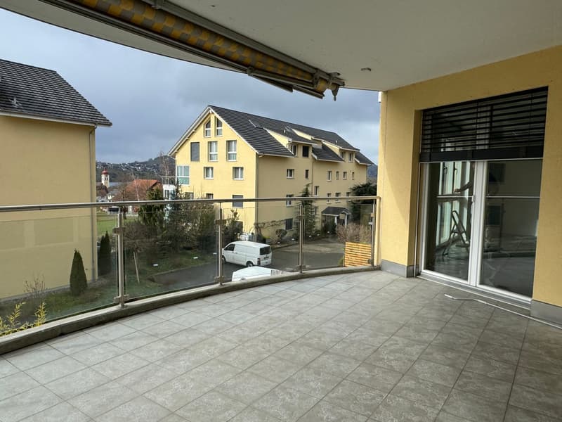 Balcony with a view of the town, yellow building, metal railings, tiled floor, open view of another house and road.