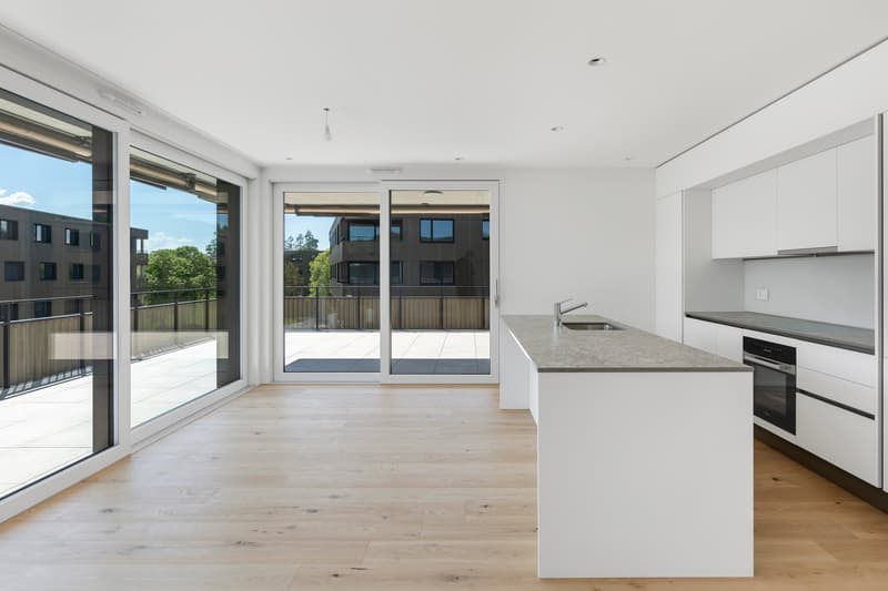 Spacious open-plan living area with large windows and sliding glass doors leading to a balcony, hardwood floors, modern kitchen with white cabinets and countertops, and a view of the surrounding buildings.