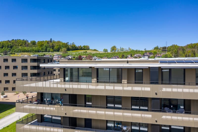 Multi-story building with a modern, modular design featuring large windows and balconies. The building has a mix of glass and wood-paneled exterior. The surrounding landscape includes green hills and trees.