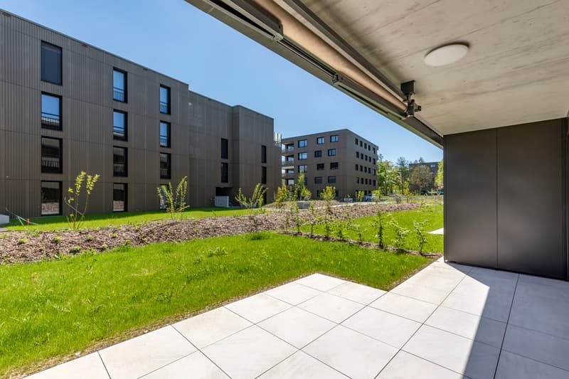 Multi-story apartment building with a modern, minimalist design featuring dark gray exterior walls and large windows. The building is surrounded by a well-maintained landscaped area with a grassy lawn, newly planted trees, and a paved walkway leading to t