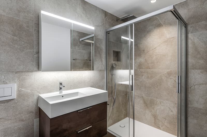 Modern bathroom with gray marble-like tiles, a white sink vanity, a large mirror cabinet, and a glass-enclosed shower area.