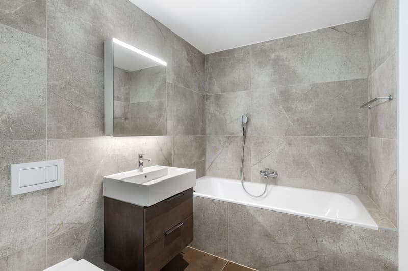Large bathroom with grey marble-like tiles covering the walls and floor. The bathroom features a white sink vanity with drawers, a large mirror, and a bathtub with a shower head. The room is well-lit with recessed lighting.