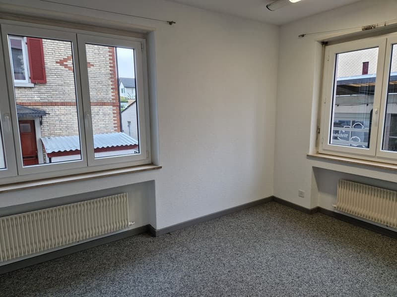 Empty room with white walls, gray floor tiles, and large windows overlooking a brick building exterior.