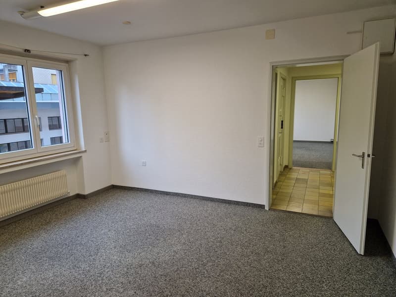 Empty room with white walls, ceiling-mounted light, radiator below the window, carpeted floor, open door leading to another room with tiled floor.