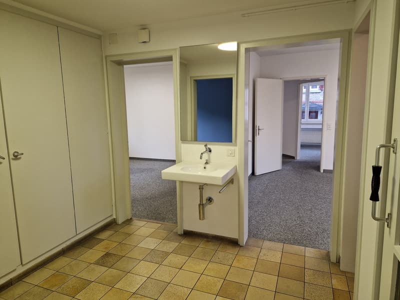 Square sink, mirror, tiled flooring, gray carpeted hallway, white doors, power outlet, silver faucet