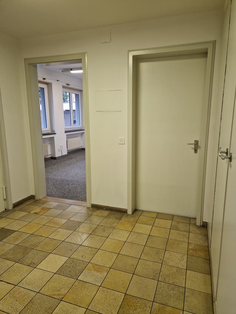 The image shows a hallway with a tiled floor and white walls. There are two doors visible, one leading to another room. The hallway appears to be in a residential or office building.