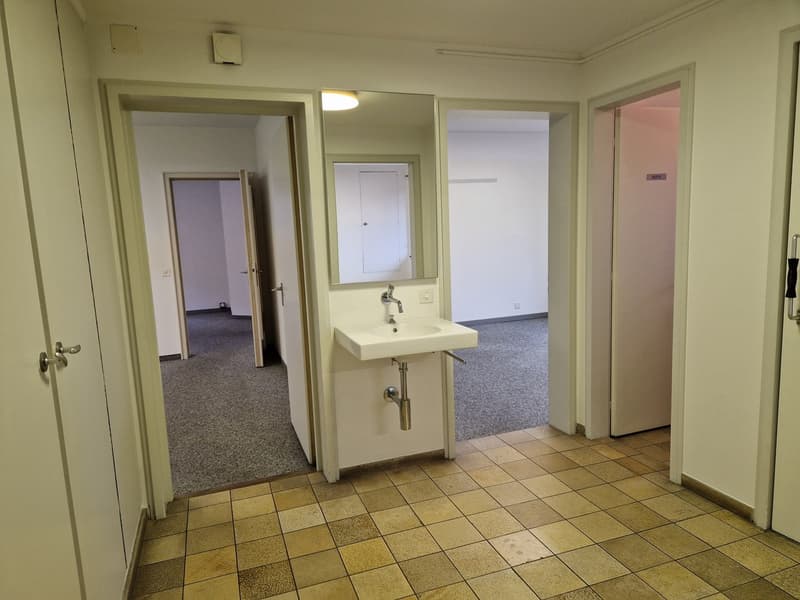 The image shows the interior of an apartment. The main room has a tiled floor and a sink with a mirror mounted on the wall. There are multiple doorways leading to other rooms. The overall appearance is clean and minimalist.