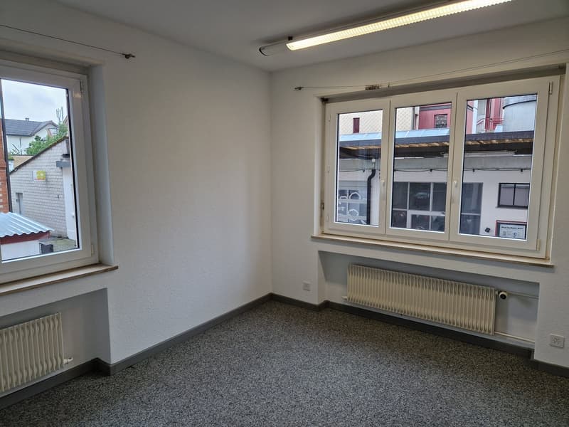 The image shows a small office space with white walls, a large window, and a radiator. The floor is covered in a gray speckled carpet. The room appears to be empty and ready for use.