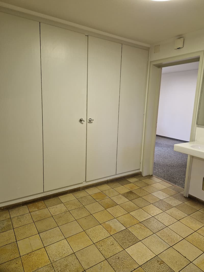 The image shows a room with white walls, a tiled floor, and built-in closets or cabinets along one wall. The room appears to be a bedroom or living space, with a doorway visible leading to another room.