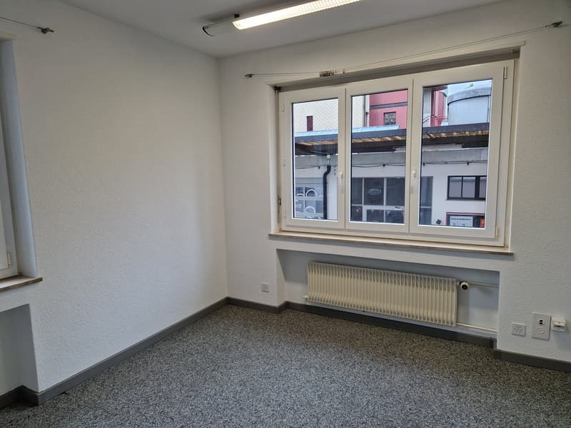 The image shows an empty office space with white walls, a large window, and a radiator along the wall. The floor is covered in a gray speckled carpet. The room appears to be a simple, functional office space.
