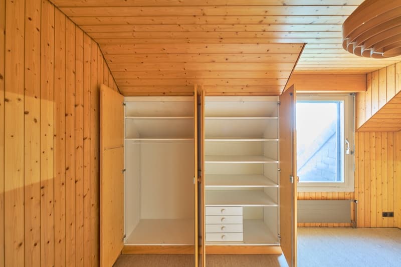 The image shows a bedroom with a wooden interior. The room has built-in closets and shelves along the walls, providing ample storage space. The ceiling is also made of wood, creating a cozy and rustic atmosphere. There is a window that allows natural ligh