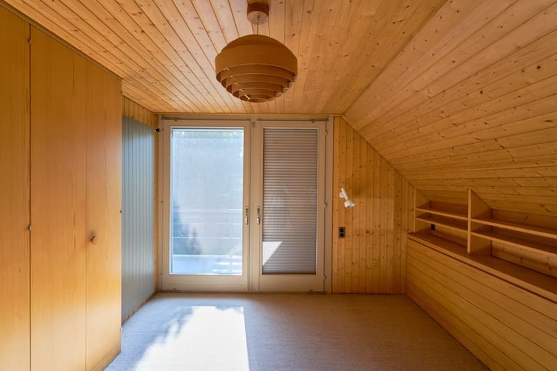 Wooden paneled room with a vaulted ceiling, large windows, and a wooden shelving unit. The room has a wooden floor and a circular pendant light fixture.