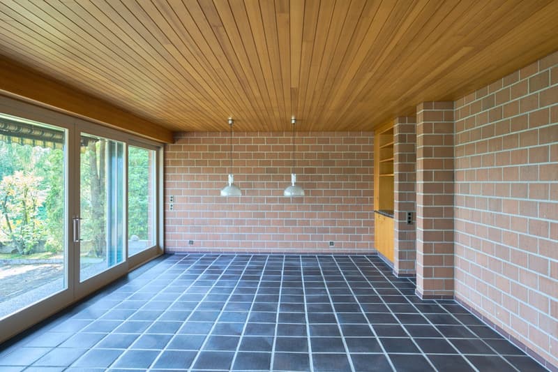 Large open space with wooden ceiling, brick walls, and tiled flooring. The room has large windows overlooking the outdoor area.