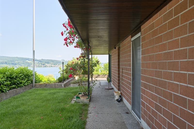 Brick exterior, covered patio, lush landscaping with rose bushes, view of a lake or body of water in the background