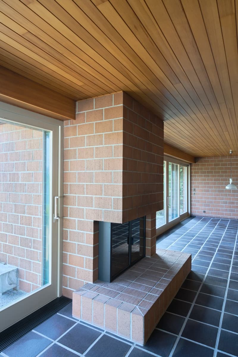 The image shows a covered patio or sunroom with a brick fireplace and a wooden ceiling with diagonal slats. The floor is tiled in a dark color. There are large windows and sliding glass doors that provide access to the outdoor space.