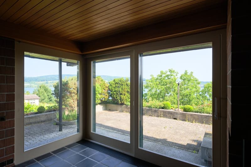 This image shows a room with large windows overlooking a scenic outdoor area. The room has a wooden ceiling and tile flooring. Through the windows, a paved patio or terrace can be seen, surrounded by lush greenery and trees.