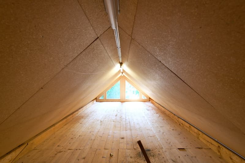 The image shows the interior of an attic space. The room has a triangular shape with a peaked ceiling and wooden floors. There is a window at the far end, allowing natural light to enter the space. The room appears to be unfinished, with exposed wooden be