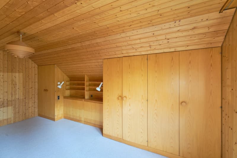 This image shows a room with a wooden ceiling and walls. The room has built-in wooden cabinets and shelves along one wall. The floor is covered with a blue carpet. The room appears to be a storage or utility space based on the simple furnishings and layou