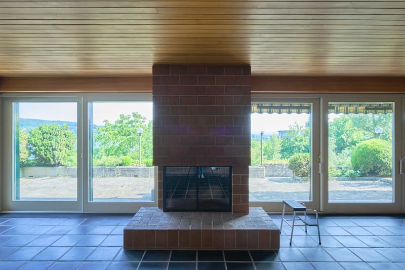 Large windows with a view of the lush greenery outside, a brick fireplace in the center, and a tiled floor.