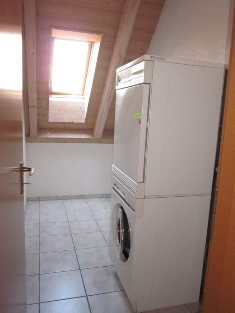 Small room with white tiled floor, wooden ceiling, a washing machine, and a dryer stacked on top of each other, a door, and a window.