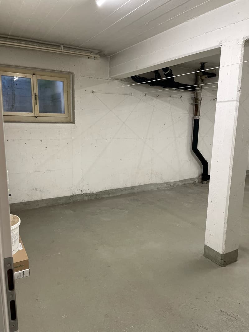 Empty room with white walls, concrete floor, small window, pipes on the ceiling, and a bucket on the floor