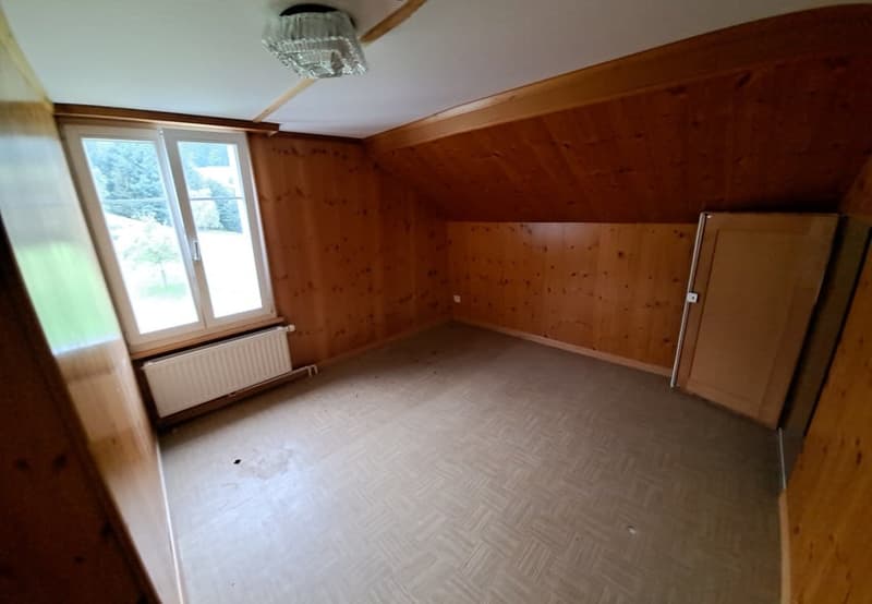 The image shows a spacious room with wooden walls and a slanted ceiling. The room has a large window that provides natural light. There is a refrigerator or other appliance visible in the corner of the room.