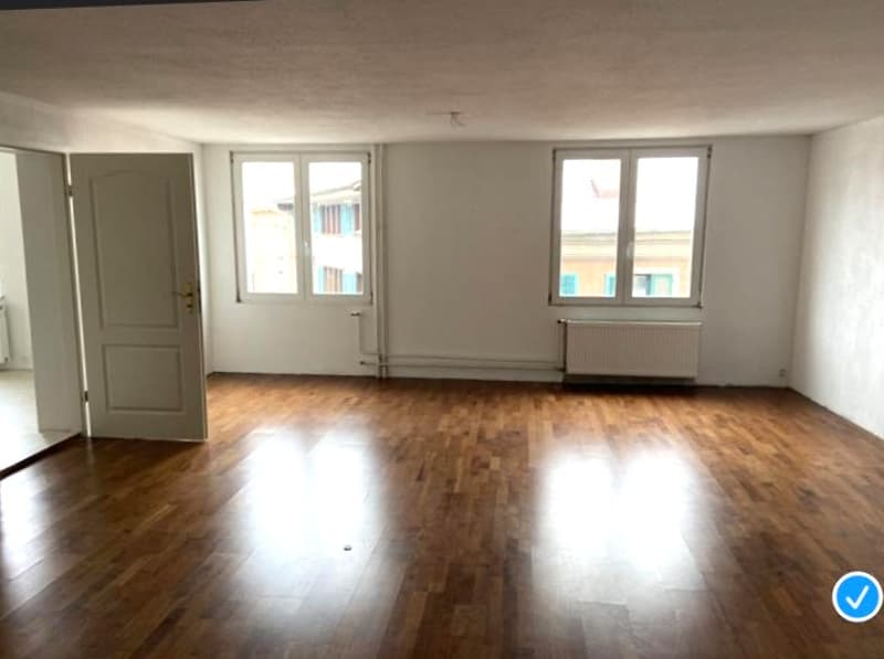Hardwood floor, white walls, large windows, radiator, open door.