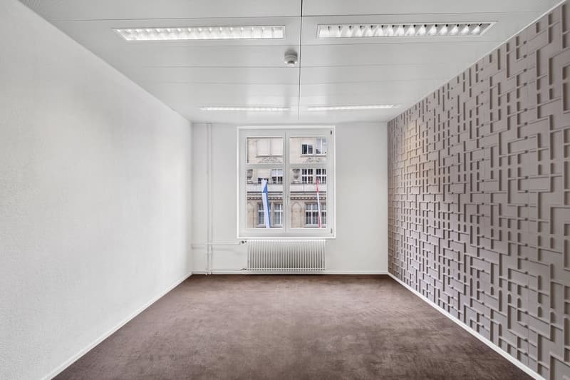 This image shows an empty office space with white walls, a large window, and a textured accent wall. The room has a modern, minimalist design with recessed lighting fixtures in the ceiling. The floor is covered in a neutral-colored carpet. Overall, the sp