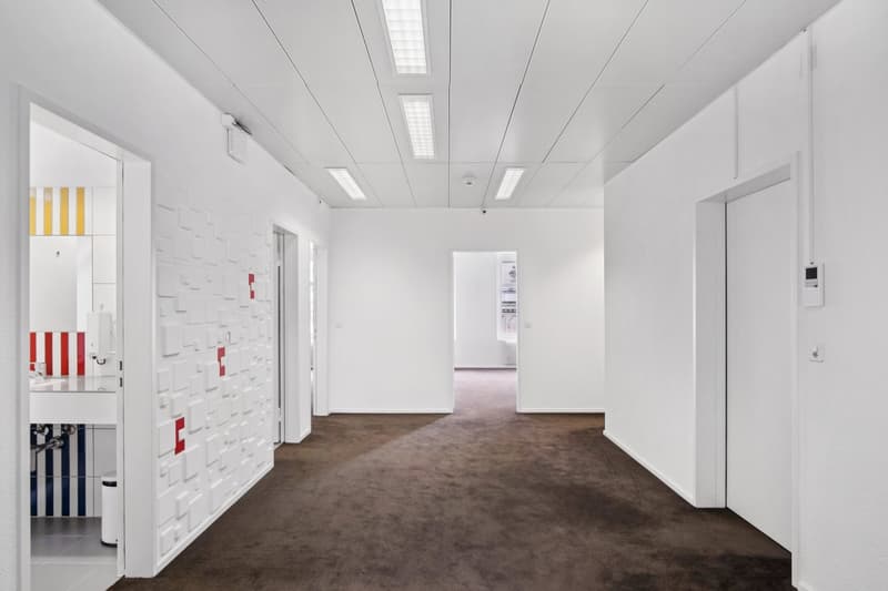 The image shows a spacious, minimalist office space with white walls, a suspended ceiling with recessed lighting, and a brown carpeted floor. The room appears to be empty, with no furniture or decor visible. There is a small storage unit or shelving unit