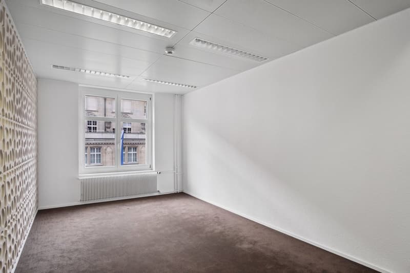 The image shows a spacious, well-lit office space with white walls, a tiled ceiling, and large windows overlooking a city street. The room has a neutral, minimalist design with a gray carpeted floor. The space appears to be suitable for use as an office o