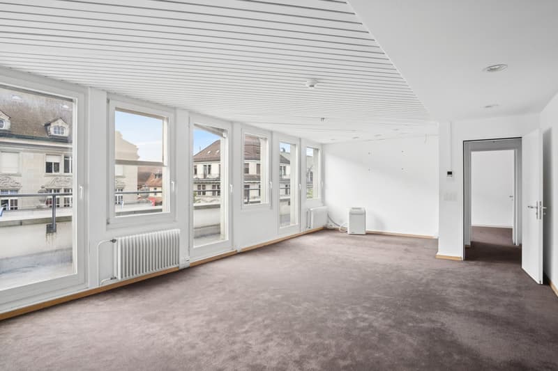 Large open-plan room with floor-to-ceiling windows offering a view of the surrounding buildings, hardwood floors, white walls, and a radiator heater.