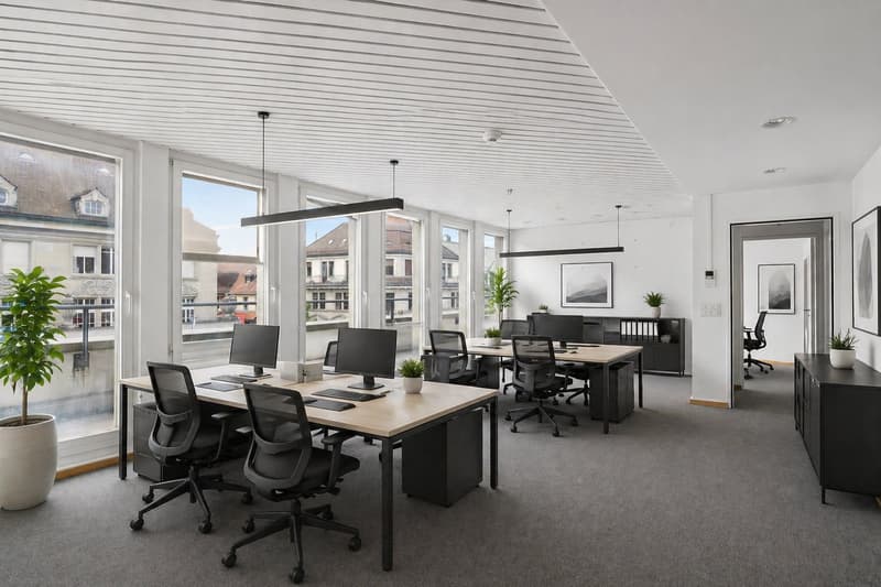 Modern office space with desks, chairs, and computer monitors. Potted plants and framed pictures on the wall.