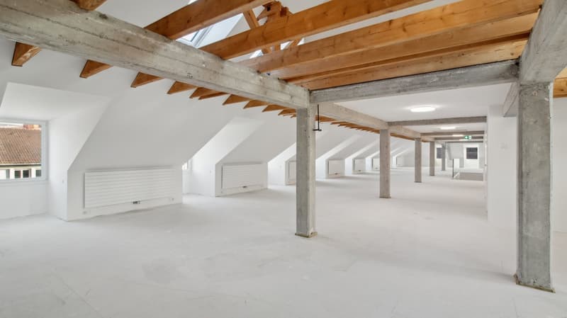A large empty room with white walls and ceiling, wooden beams, metal pillars, and several windows. The room is vacant, and there is a light on the ceiling.