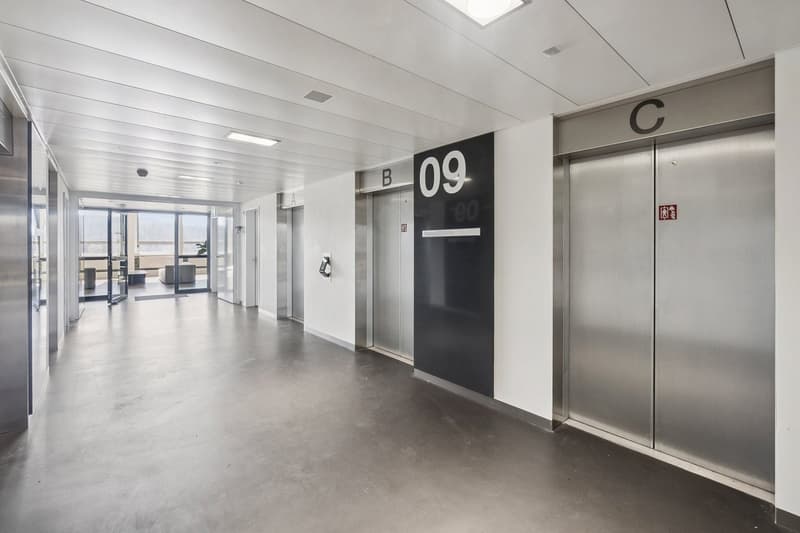 Empty hallway with three elevators, labeled A, B, and C. The floor is gray, and the walls are white. There are lights on the ceiling.