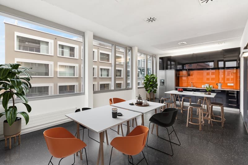 modern office room with large windows, white tables, orange chairs, plants, kitchen area with orange wall