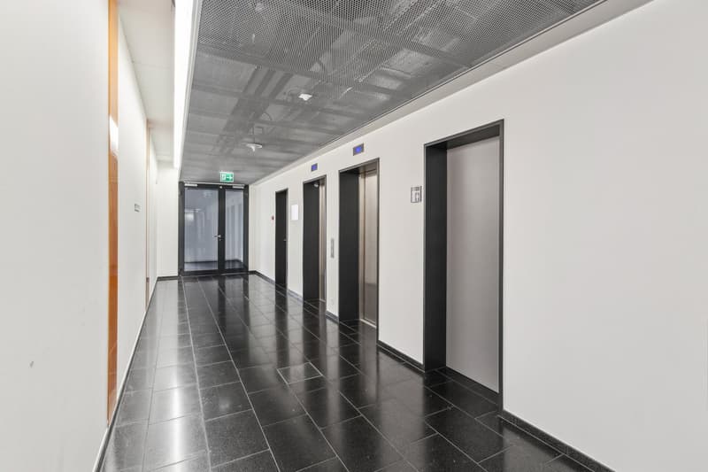 Empty hallway with black tiles, white walls, and metal ceiling. Three elevator doors are visible.
