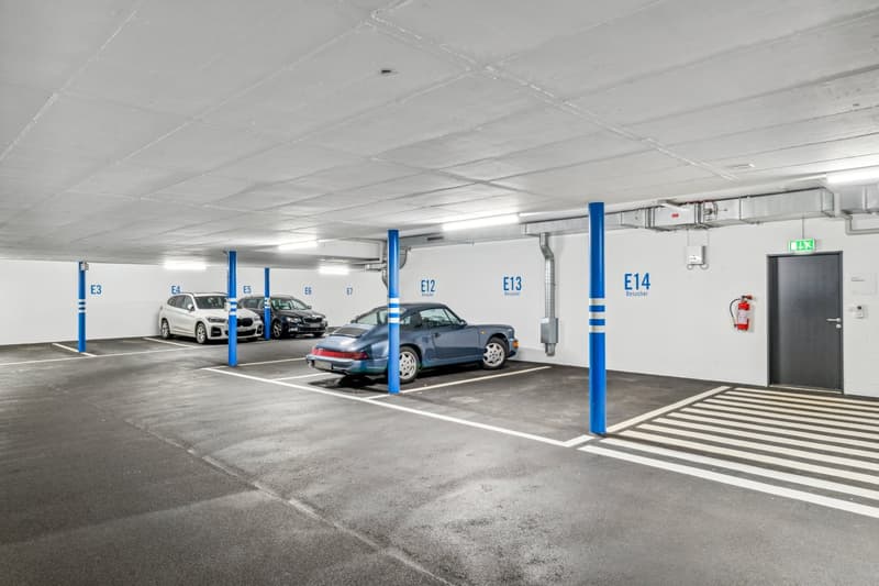 Underground parking area with numbered parking spots, blue poles, white walls, and a fire extinguisher