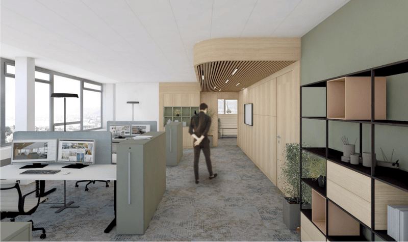 Open office space with desks, monitors, chairs, and a person walking. Wooden and green walls, ceiling with lights, and a shelf with plants.