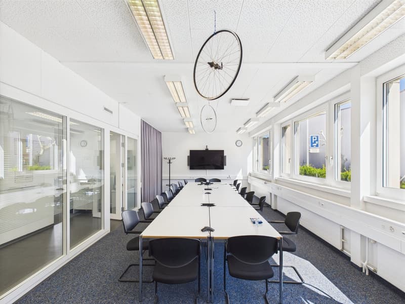 Large conference room with a long wooden table, black chairs, a TV mounted on the wall, and bicycle wheels hanging from the ceiling. The room has large windows and a bright, modern design.