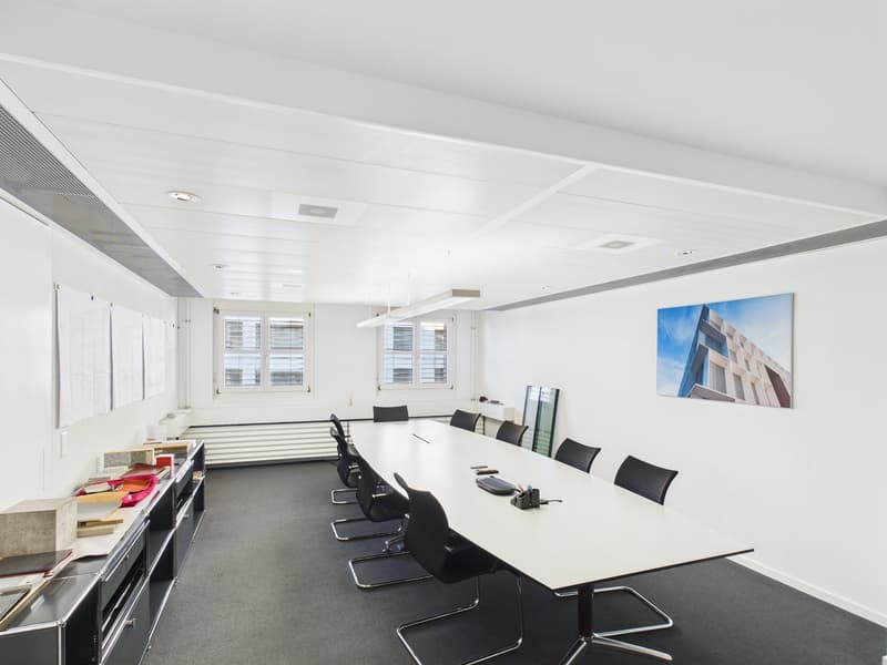 A rectangular office room with white walls, black chairs, a long white table, grey carpet, two large windows, picture and objects on the table