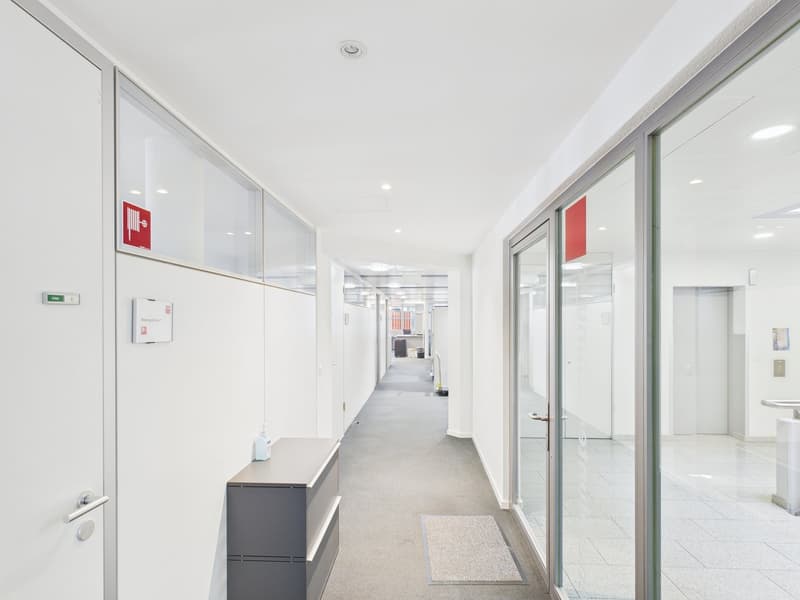 modern office corridor with white walls, carpeted floor, glass partitions, fire extinguisher sign, modern furnishings, and a small table with a bottle