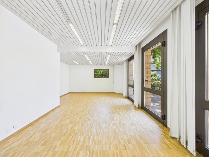 This image shows a large, open room with a high, vaulted ceiling and white paneled walls. The floor is made of light-colored hardwood or laminate flooring. There is a large window at the far end of the room, providing natural light and a view of the outdo