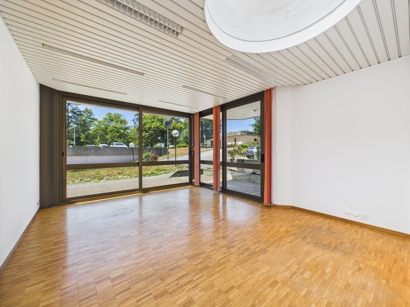 Large open space with hardwood flooring, white walls, and large windows providing a view of the outdoor area. The room has a high ceiling with a circular light fixture.