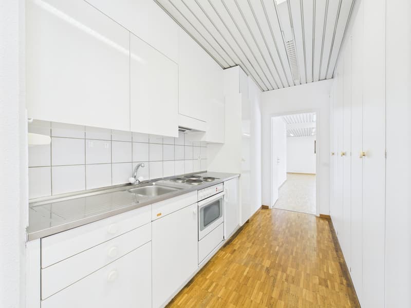 Bright and modern kitchen with white cabinets, stainless steel appliances including a dishwasher, and a tiled backsplash. The kitchen leads to a hallway with white walls and a wooden floor.