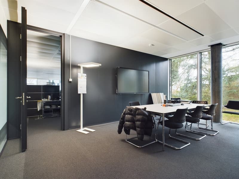 Modern office space with black walls, large windows, a TV mounted on the wall, a conference table with black chairs, and a kitchenette area visible through the open doorway.