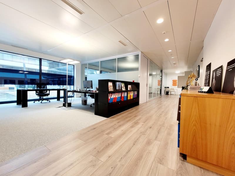 Spacious open-plan office with hardwood flooring, glass partitions, desks, computers, and storage units. The office has a modern and professional design with good natural lighting from the large windows.